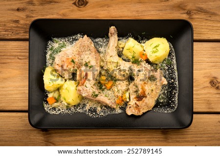 Rabbit cooked in sour cream with vegetables on the black plate on the wooden background.