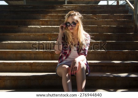 Trendy hipster girl sitting on stairs, looking over sunglasses. Youth street style.