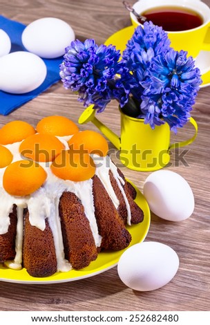 Easter cake with apricots on a yellow plate on a wooden background with eggs and blue flowers in a yellow watering can