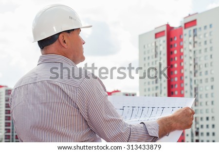 An engineer in white helmet examining blueprints on a background with buildings