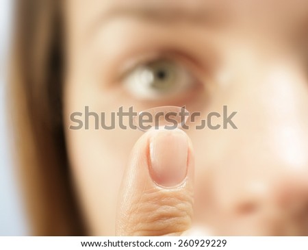 HOLDING UP A CONTACT LENS - A close up of a contact lens on a finger with an out of focus eye in the background