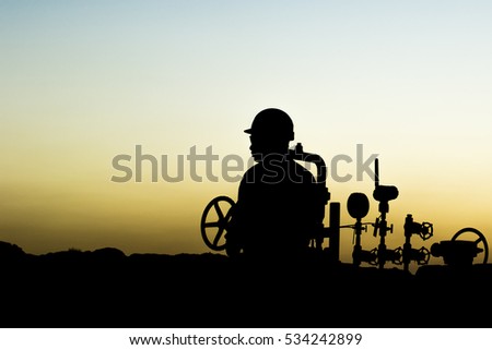 Silhouette of oilfield worker monitoring valves at sunset.