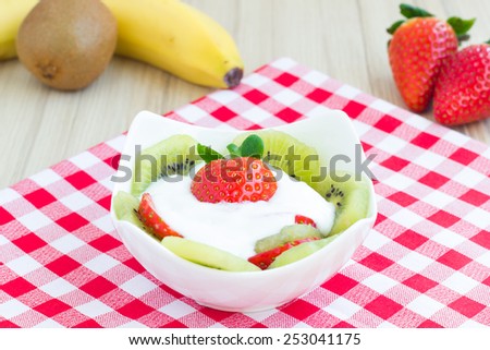 Fruit salad with kiwi, banana, strawberry and yogurt sauce on a red checkered table cloth.