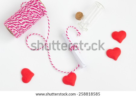 Message in a bottle. Roll of white paper with tiny glass, pink thread and red hearts on a white background. Up view.