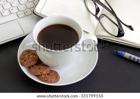 A cup of coffee with cookies, laptop, an open book and glasses on a desk.