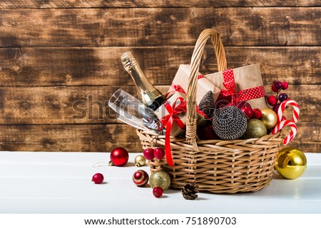 Christmas basket with champagne bottle and glasses, gifts with red satin ribbon, candy canes, pine cones, golden garlands on dark brown wooden background