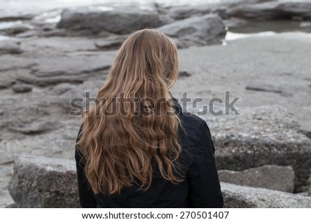 Woman with long blond hair sitting along on stones with her back to the camera