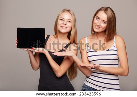 Two girls showing tablet computer screen , smiling, on a gray background