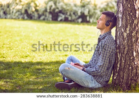Music, technology, people and leisure concept - close up of teenage boy in headphones with book listening to music online at park