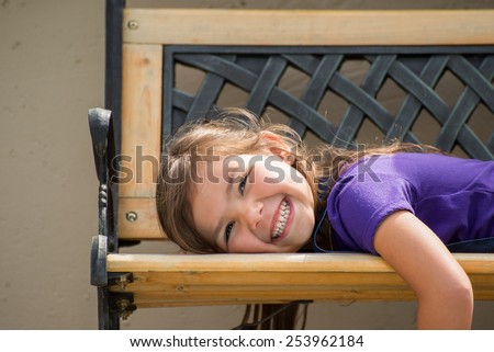 Little girl lying on her stomach on an outside bench with a big smile on her face.