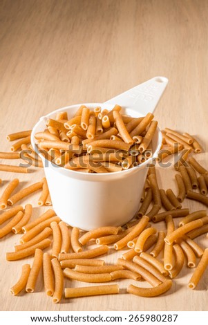 A standard cup size measuring cup of white plastic sits on a wooden surface, filled to the brim with wholewheat macaroni, while much more macaroni lies spilled around the cup.