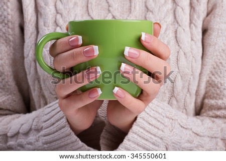 Woman holds a green cup close up. Woman hands with elegant french manicure nails design holding a cozy knitted mug. Knitted winter sweater.