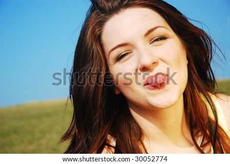 A beautiful young woman sticking her tongue out on a field with a blue sky.