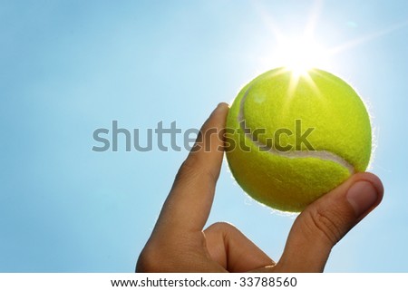 A man\'s hand holding a tennis ball up to a blue sky with the sun behind it.