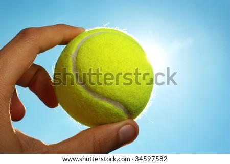 A man\'s hand holding a tennis ball up to a blue sky with the sun behind it.