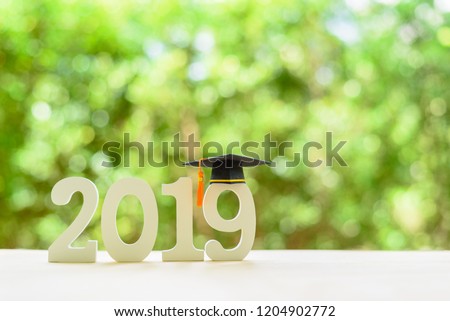 2019 happy new year / graduate study abroad program, time schedule arrangement, education concept : Black graduation cap of success on number 2019 white wood cut on desk table, nature green background