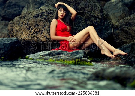 brunette lady in wet red dress sitting on the stone in the sea water