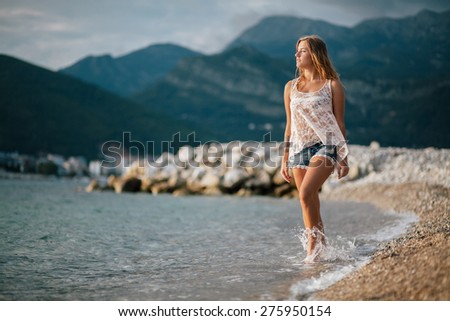 dreamy fashion girl in creamy blouse and blue jeans shirts walk on beach with mountains background in Montenegro