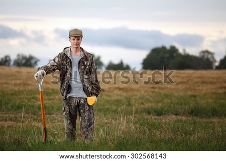 Man with metal detector and shovel posing in the field. Treasure hunter is wearing camouflage and has confident look