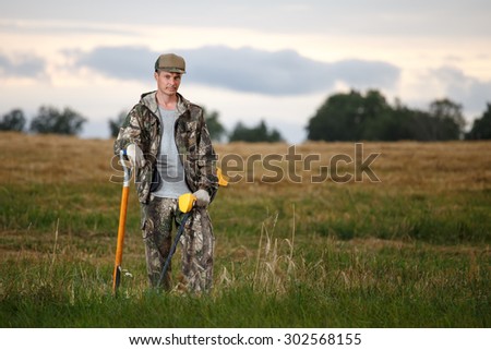 Treasure hunter with metal detector leaned on a shovel in field. Confident look and camouflage clothes. Rural background