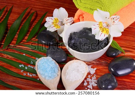 black therapeutic Spa treatments clay in a bowl on a red wooden table flowers salt sea two wooden spoons stones for massage and two towels