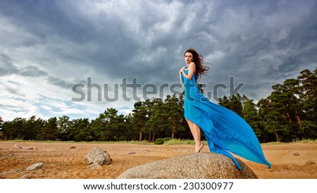 Slim girl in blue flowing dress against the sky stands on the stone