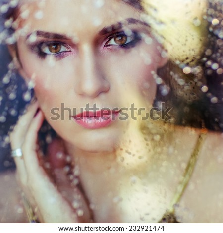 very beautiful brunette with brown eyes behind the glass, through which drain water drops, close up