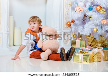 Small very cute blond boy sitting by the fireplace and white Christmas tree