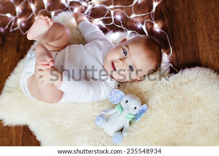 Very cute plump baby lying on the floor on the background of Christmas lights next to the soft toy and she holds her legs