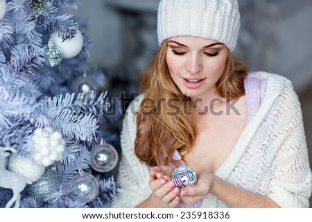 Very beautiful red-haired girl with blue eyes in a white hat about silver Christmas tree and looks at the ball
