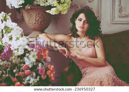 Very beautiful girl brunette in pink dress sitting on the couch surrounded by flowers