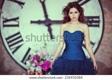 Very beautiful sexy brunette girl in a blue dress standing amid giant hours with flowing hair