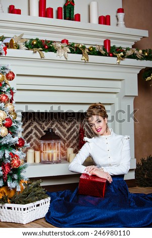 Beautiful blonde girl in a blue skirt sitting near the Christmas tree and fireplace