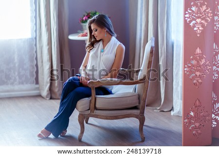 Very beautiful pregnant girl with gorgeous hair in a white blouse and pants sitting in the chair against the window profile