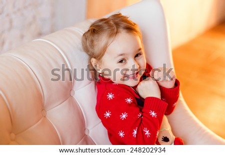 Little girl child with brown eyes smiles provocatively, in a red sweater on the background of yellow lights