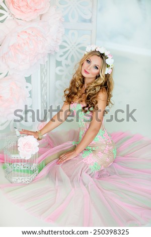 Very beautiful sensual girl with curly blond hair and a wreath of delicate spring flowers, sitting on the background of large pink flowers and white cells