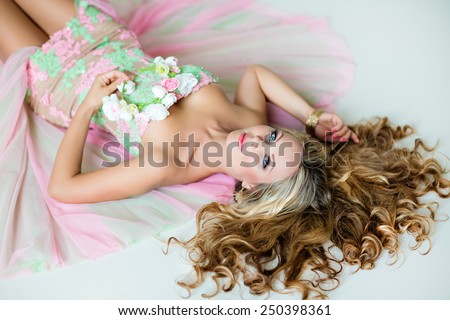 Very beautiful sexy sensual girl with curly blond hair, blue eyes in a lace dress and a wreath of delicate spring flowers, lying on white floor