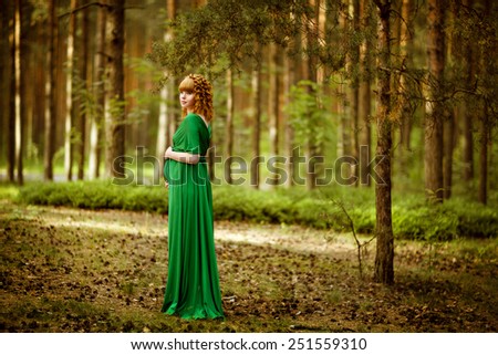 Portrait of a beautiful curly haired pregnant girl in a green dress, standing thoughtfully in the woods among the pines