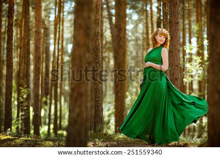 Portrait of a beautiful curly haired pregnant girl in a green flowing dress in the woods among the pines