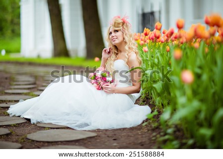 Very beautiful bride blonde in a white dress with an unusual stylish hairstyle in the form of a hat sits about tulips and smiles