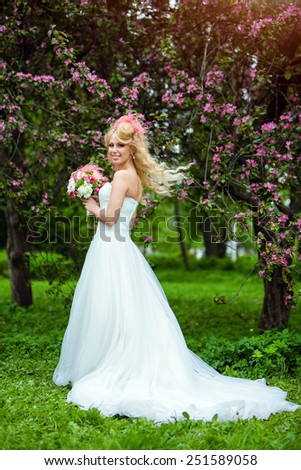 Very beautiful happy bride blonde curly hair in a white dress and unusual bouquet in hand, against the background of Apple blossoms in full growth