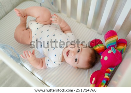 Very cute little baby boy lying on the bed at the back in the crib next to a bright toy
