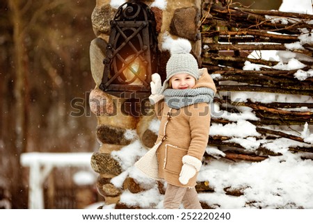 Very sweet beautiful little girl child in a beige coat defiantly smiling against winter village near the lantern