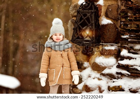 Very sweet beautiful little girl child in the beige coat is on the background of the winter village near the lantern