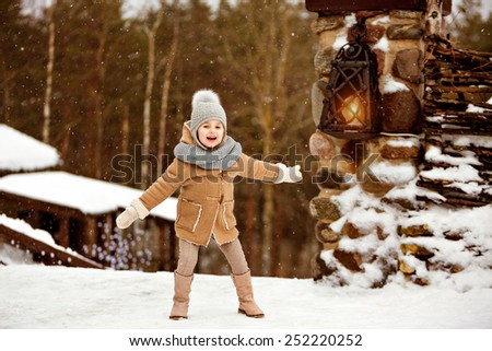 Very sweet beautiful little girl child in the beige coat is happy and smiling against winter village near the lantern