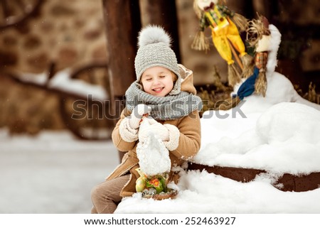 Very sweet and funny girl child in a beige coat sitting on the snow and holding a chicken