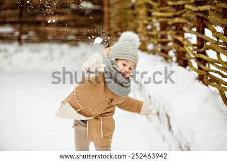 Very nice beautiful girl child in a beige coat and a gray hat throws snowballs against a wooden gate in winter