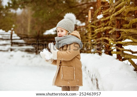 Very nice beautiful girl child in a beige coat and a gray hat sculpts snowballs against a wooden gate in winter