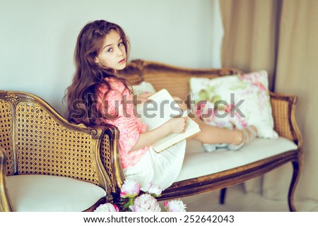 Cute sad girl baby with gorgeous hair in the skirt and the sweater is reading a book, sitting on the sofa ,in the interior