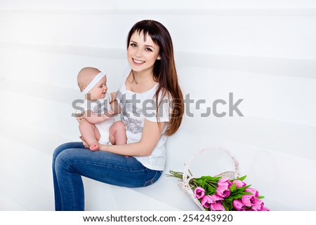 Beautiful and happy mom brunette in white t-shirt and jeans holding a little smiling baby daughter on a white background with tulips